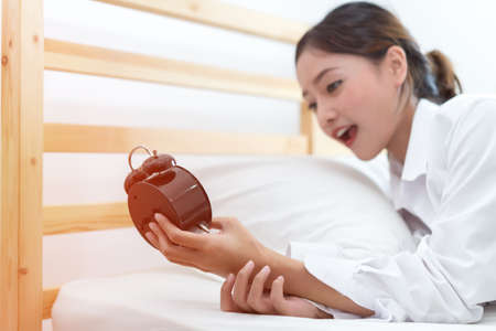 Woman wake up late in the morning looking alarm clock snooze or turn off under blanket on bed at roomの写真素材