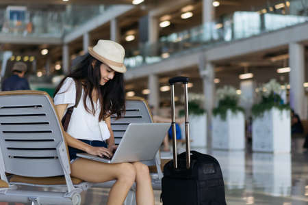 Asian woman teenager using laptop computer at airport terminal sitting with luggage suitcase and backpack for travel in vacation summer relaxing waiting flight transportの写真素材