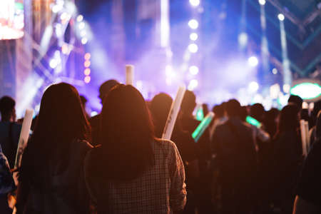Two women friends crowd concert stage lights and people fan audience silhouette raising hands glow stick in the music festival rear view with spotlight glowing effectの写真素材