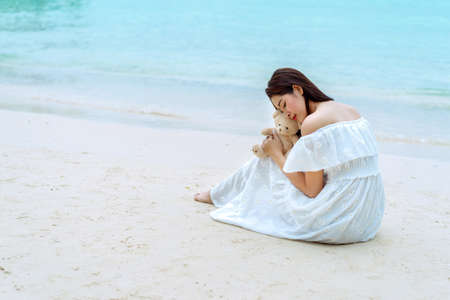 Asian single woman sitting with teddy bear at seaside beach wearing white dressの写真素材