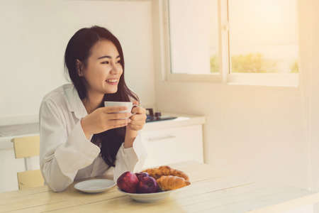 Asian woman drinking coffee in the morning and eating bread,apple fruit healthy food breakfast meal in kitchen room fresh start the day at home healthy lifestyle conceptの写真素材