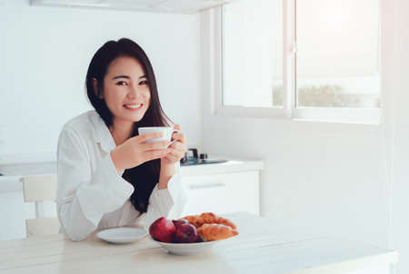 Asian woman drinking coffee in the morning and eating bread,apple fruit healthy food breakfast meal in kitchen room fresh start the day at home healthy lifestyle conceptの写真素材