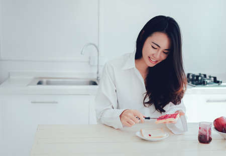 Asian woman slice jam bread in the morning and eating apple fruit,coffee healthy food breakfast meal in kitchen room fresh start the day at home healthy lifestyle conceptの写真素材