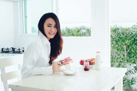 Asian woman slice jam bread in the morning and eating apple fruit,coffee healthy food breakfast meal in kitchen room fresh start the day at home healthy lifestyle conceptの写真素材