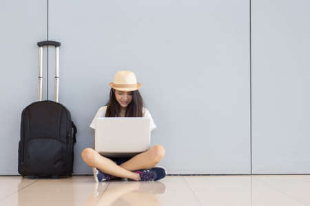 Asian woman teenager using laptop computer at airport terminal sitting with luggage suitcase and backpack for travel in vacation summer relaxing waiting flight transport or booking ticket and online check inの写真素材