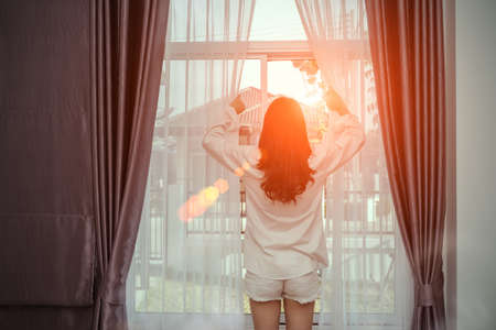 Young woman wake up in the morning and open window after waking on bed relaxing good day in holiday with sunlight, rear view at living home lifestyleの写真素材