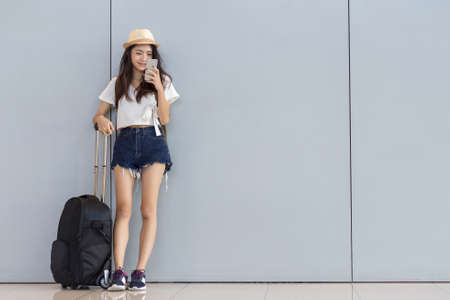 Asian woman teenager using smartphone at airport terminal standing with luggage suitcase and backpack for travel in vacation summer relaxing waiting flight transport online check in or booking ticketの写真素材