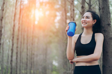 Beautiful asian woman drinking protein shake in the forest nature for relaxing after runner exercise cardio workout or loss of sweat recovery refreshment active healthy lifestyle at mountain hill.の写真素材