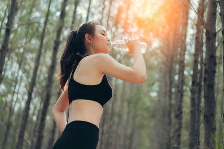Beautiful asian woman drinking cool water bottle in the spruce forest nature for relaxing after runner exercise cardio workout or loss of sweat recovery refreshment active healthy lifestyle.の写真素材