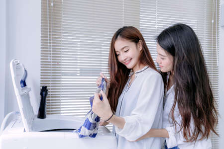 Same sex couple women asian doing housework or chores helping embracing with washing machine laundry loading clothes and in the room, Lesbian relationship female living together.の写真素材