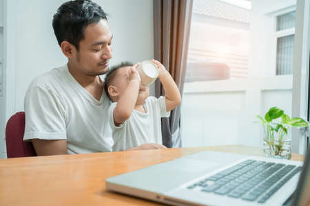 Asian father teaching little son drinking milk for calcium and protein nutrition at morning together on workplace at home with laptop computer.の写真素材