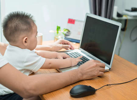 Asian father and little son typing something on laptop computer on workplace at office home...