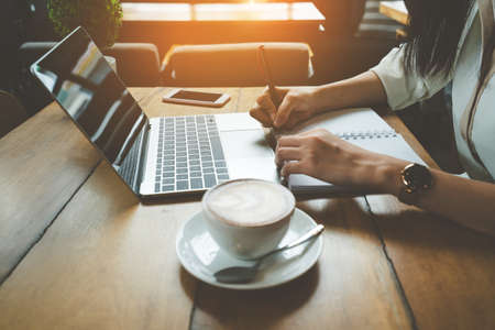 Freelancer asian woman working job and writing note with digital laptop computer drinking coffee breakfast on workplace table at cafe shop.の写真素材