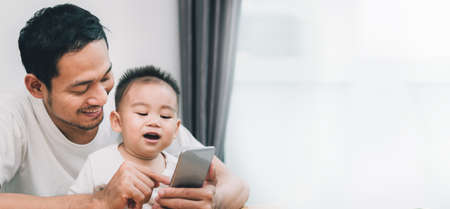 Asian father and little son watching video from smartphone at home happiness laughing together, happy family concept of panoramic copy space the right web banner.の写真素材