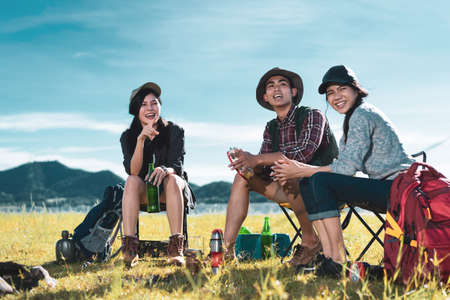Camping group of friends asian people together, teenagers travel with backpack,tent man playing song relaxing on vacation time holiday sitting on chairs near river and mountain view.の写真素材
