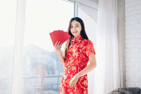 Chinese New Year of Portrait Beauty Asian Woman with Chinese dress,Qipao,Cheongsam Smile holding Red envelope Standing near Window in Clean White Room,Copy Space.の写真素材