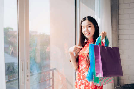 Shopping Asian Woman Chinese dress,Qipao,Cheongsam Chinese New Year and hold Bag Sitting near Window in Clean White Room,Copy Space.の写真素材