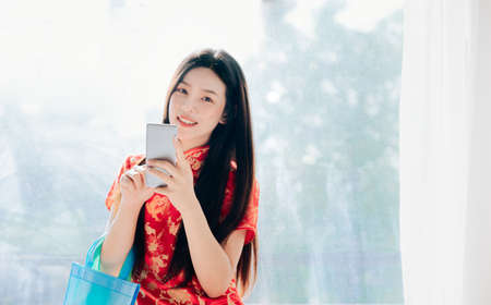Beauty Asian Woman with Chinese dress,Qipao,Cheongsam Chinese New Year Using Smartphone Technology Shopping Online Standing near Window in Clean White Room,Copy Space.の写真素材