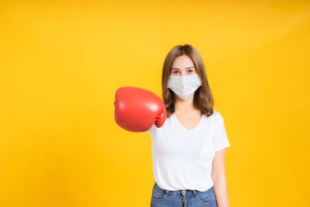 Young asian woman with medical mask protection coronavirus COVID-19 fighting wearing boxing gloves healthy and safety care concept on yellow background isolated studio shot, copy space.の写真素材