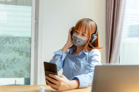 Asian woman wearing mask staying at home listening music with smartphone, headphone relaxing...