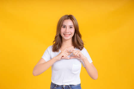Portrait happy young asian woman laughing finger hands heart gesture romantic love joyful funny positive emotion in white t-shirt, Yellow background isolated studio shot and copy space.の写真素材
