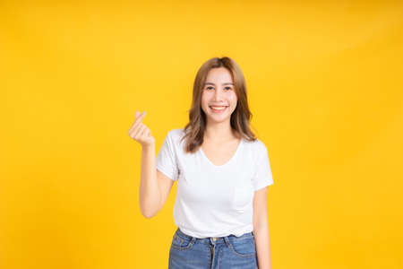 Portrait happy young asian woman laughing finger hands heart gesture romantic love joyful funny positive emotion in white t-shirt, Yellow background isolated studio shot and copy space.の写真素材