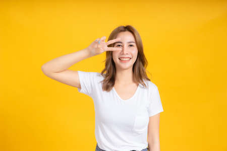 Portrait happy young asian woman laughing finger hands v-symbol gesture selfie joyful funny positive emotion in white t-shirt, Yellow background isolated studio shot and copy space.の写真素材