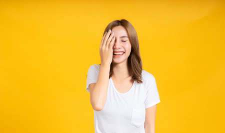 Portrait young happy asian woman smile laughing hand covering your eye standing enjoy fun relaxing carefree positive emotion in white t-shirt, Yellow background isolated studio shot and copy space.の写真素材