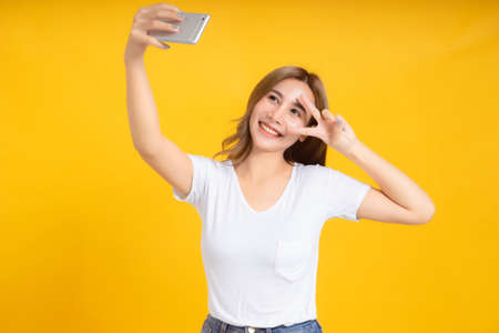 Portrait happy young asian woman laughing finger hands v-sign eye gesture selfie joyful funny positive emotion in white t-shirt, Yellow background isolated studio shot and copy space.の写真素材