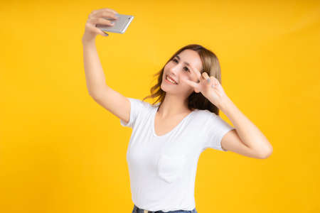 Portrait happy young asian woman laughing finger hands v-sign eye gesture selfie joyful funny positive emotion in white t-shirt, Yellow background isolated studio shot and copy space.の写真素材