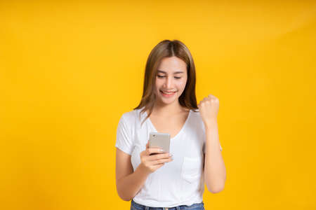 Portrait happy young asian woman glad lucky and successful with holding smartphone mobile positive emotion in white t-shirt, Yellow background isolated studio shot and copy space.の写真素材