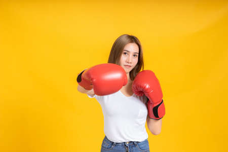 Portrait happy young asian woman with boxing glove punching hit or self defend in white t-shirt, Yellow background isolated studio shot and copy space.の写真素材