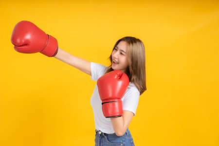 Portrait happy young asian woman with boxing glove punching hit or self defend in white t-shirt, Yellow background isolated studio shot and copy space.の写真素材