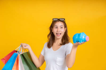 Portrait happy young asian woman holding piggybank saving money for shopping positive emotion in white t-shirt, Yellow background isolated studio shot and copy space.の写真素材
