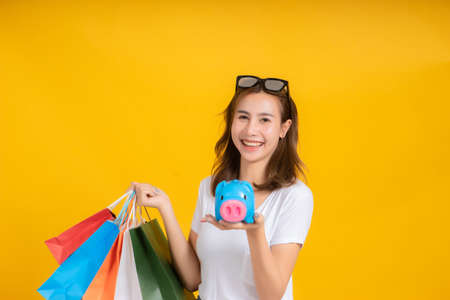 Portrait happy young asian woman holding piggybank saving money for shopping positive emotion in white t-shirt, Yellow background isolated studio shot and copy space.の写真素材
