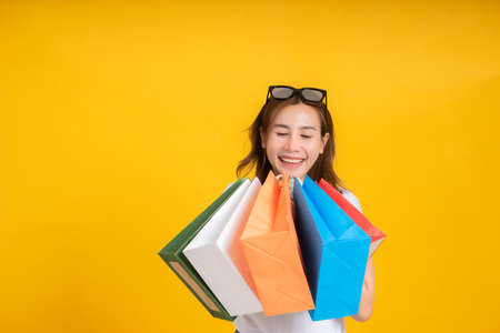 Happy young asian woman with shopping paper bag fashion purchase on store shopping mall concept in white t-shirt smiling emotion on yellow background isolated studio with copy space.の写真素材