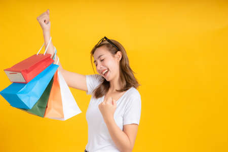 Happy young asian woman with shopping paper bag fashion purchase on store shopping mall concept in white t-shirt smiling emotion on yellow background isolated studio with copy space.の写真素材