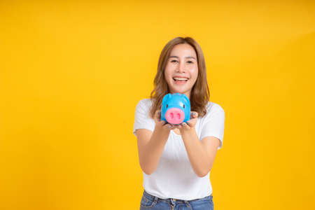 Portrait happy young asian woman holding piggybank saving money for future and business earning income positive emotion in white t-shirt, Yellow background isolated studio shot and copy space.の写真素材