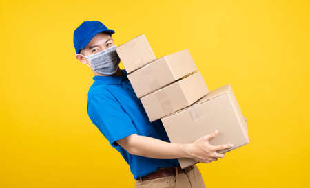 Delivery handsome man wearing mask with heavy many carton box picking up the package to front home receiver shipping deliver cargo prevention infected virus on yellow background isolated studio shot.の写真素材