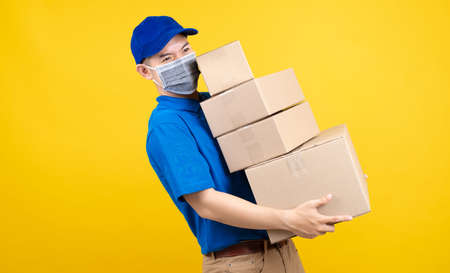 Delivery handsome man wearing mask with heavy many carton box picking up the package to front home receiver shipping deliver cargo prevention infected virus on yellow background isolated studio shot.の写真素材