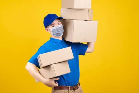 Delivery handsome man wearing mask with heavy many carton box picking up the package to front home receiver shipping deliver cargo prevention infected virus on yellow background isolated studio shot.の写真素材
