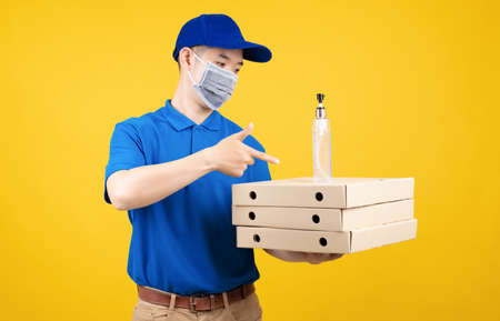 Delivery handsome man wearing mask showing hand sanitizer alcohol gel picking up the pizza food to front home receiver shipping deliver cargo prevention infected virus on yellow background isolated.の写真素材