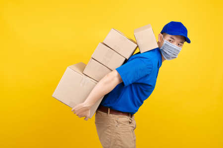 Delivery handsome man wearing mask with heavy many carton box picking up on back the package to front home receiver shipping deliver cargo prevention infected virus on yellow background isolated.の写真素材