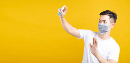 Handsome asian man wearing mask showing hand sanitizer alcohol spray for cleaning stop gesture emotion prevention infected virus crisis coronavirus, covid-19 on yellow background isolated studio shot.の写真素材