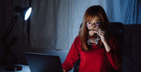 Korean woman working late time with laptop computer drinking water home office workhard overworked, Freelancer stay home business quarantine sleepless crisis coronavirus stay home dark room.の写真素材