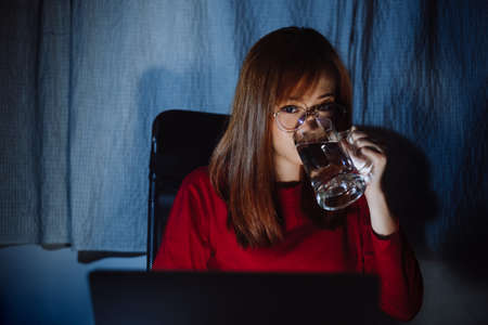 Korean woman working late time with laptop computer drinking water home office workhard overworked, Freelancer stay home business quarantine sleepless crisis coronavirus stay home dark room.の写真素材