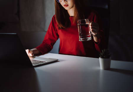 Korean woman working late time with laptop computer drinking water home office workhard overworked, Freelancer stay home business quarantine sleepless crisis coronavirus stay home dark room.の写真素材