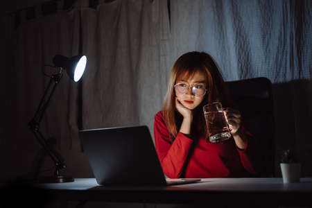 Korean woman working late time with laptop computer drinking water home office workhard overworked, Freelancer stay home business quarantine sleepless crisis coronavirus stay home dark room.の写真素材