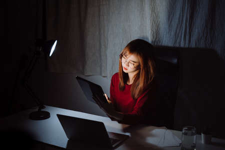 Young asian korean woman working late time with tablet, laptop computer home office work hard overworked, Freelancer stay home business quarantine sleepless crisis coronavirus stay home dark room.の写真素材