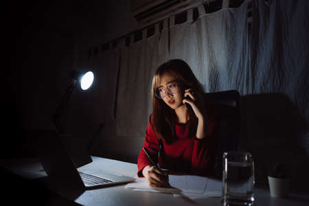 Asian woman working late time at night talking with smartphone, laptop computer home office unhappy emotion, Freelancer stay home business sleepless quarantine crisis coronavirus in dark room.の写真素材